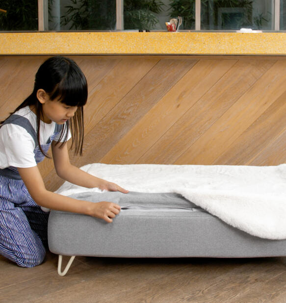 Girl zipping a white faux sheepskin topper onto Topology dog bed mattress with white hairpin feet.